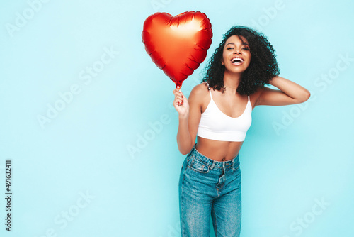 Beautiful black woman with afro curls hairstyle. Smiling model dressed in white summer clothes. Sexy carefree female posing near blue wall in studio. Tanned and cheerful. Holding heart air balloon © halayalex
