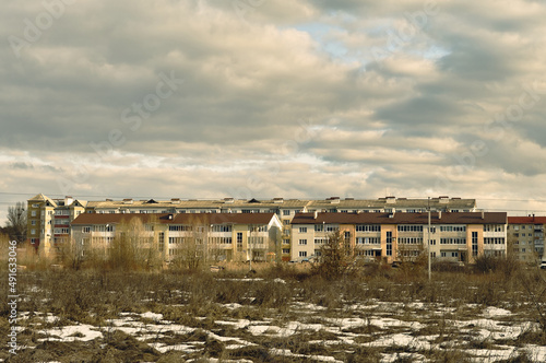 On a wasteland overgrown with dry grass, there are multi-storey buildings, the sky with clouds hangs over residential buildings, the urban landscape