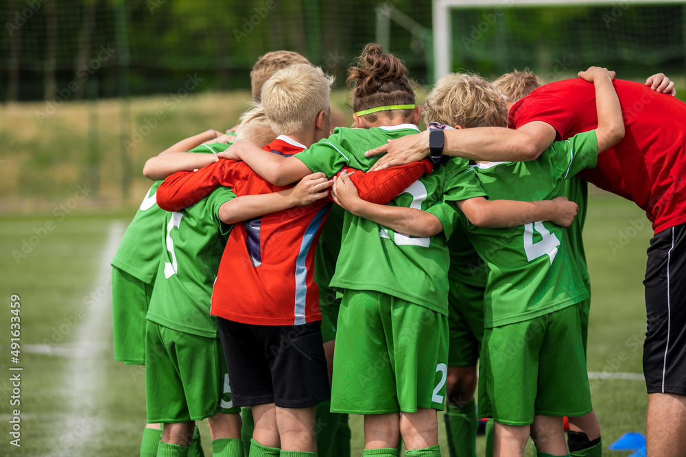 Happy sports team with a young coach. Children playing a football game on stadium pitch. Trainer motivating young players during the pregame briefing. Boys in green soccer jersey uniforms Happy sports team with a young coach. Children playing a football game on stadium pitch. Trainer motivating young players during the pregame briefing. Boys in green soccer jersey uniforms