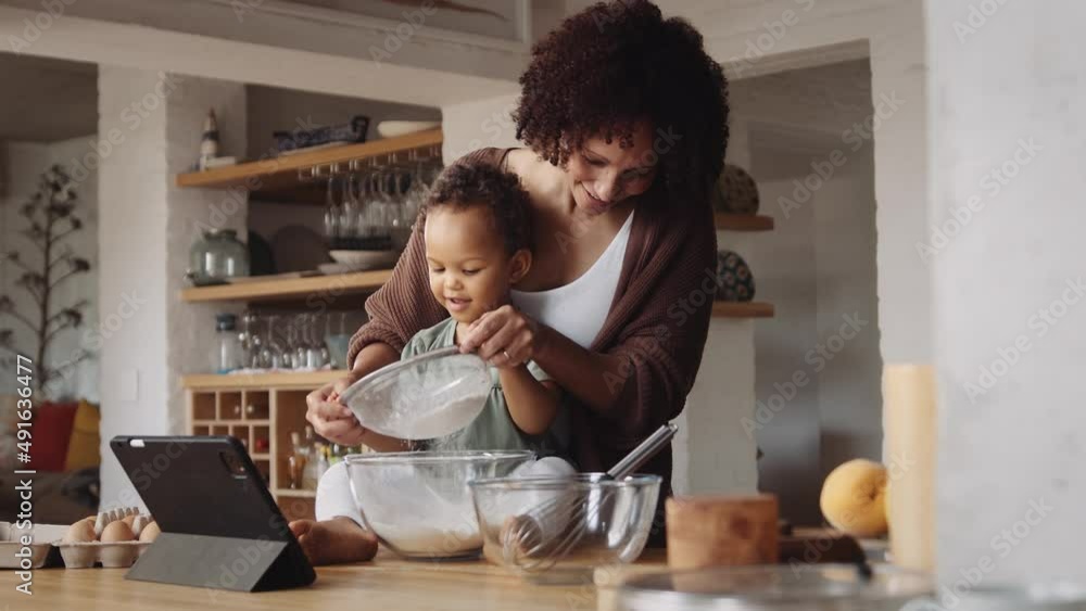 Biracial mother and daughter in modern-styled kitchen sifting flour in bowl to bake a cake. Biracial mother and daughter in modern-styled kitchen sifting flour in bowl to bake a cake.