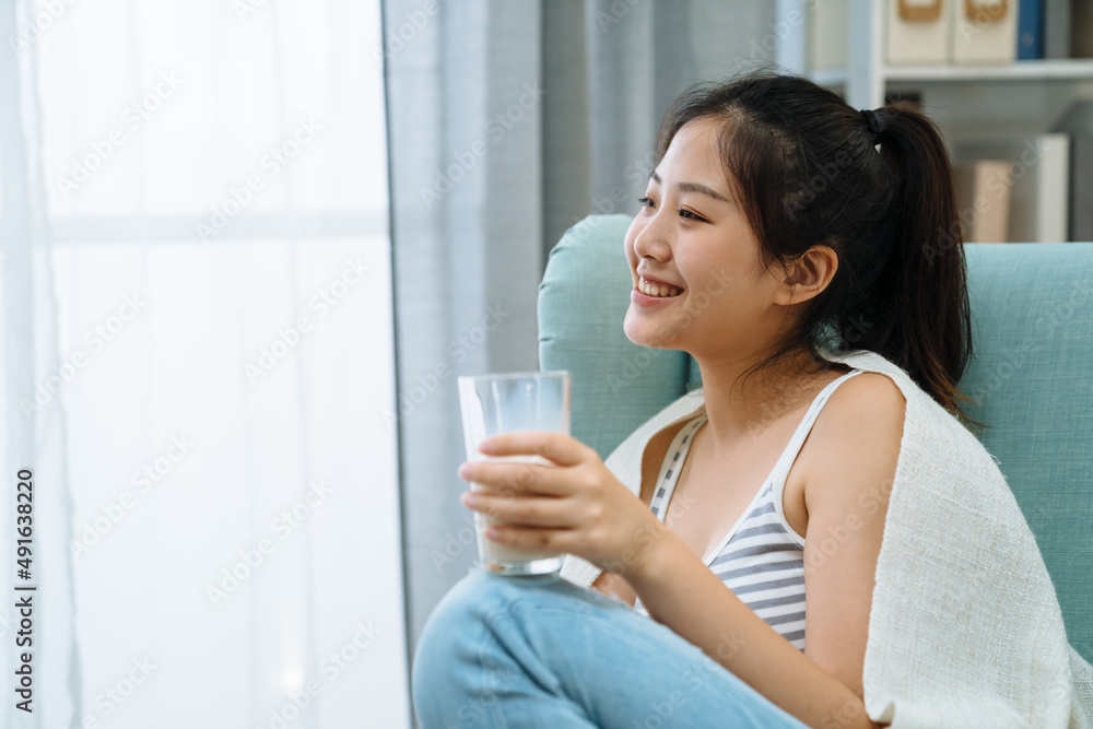 Happy young woman wrapped with warm blanket sitting on comfort sofa drinking glass of milk at home living room. charming girl enjoy breakfast beverage in single armchair indoors looking side window Happy young woman wrapped with warm blanket sitting on comfort sofa drinking glass of milk at home living room. charming girl enjoy breakfast beverage in single armchair indoors looking side window