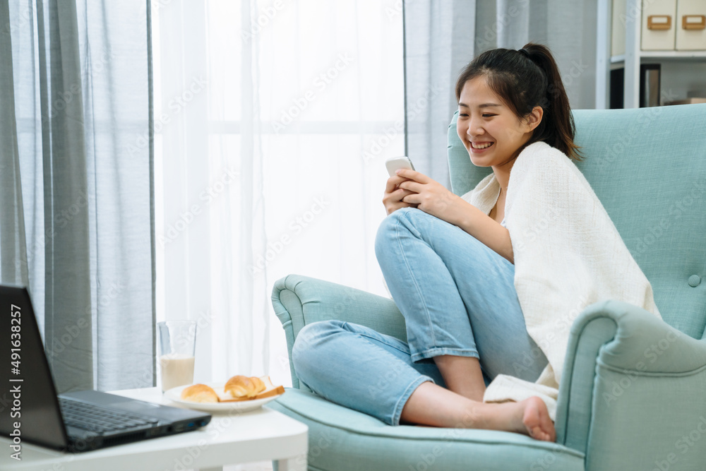 asian cute girl eating breakfast while using mobile phone and laptop computer. charming woman smiling using cellphone text message on comfort armchair by window in morning. glass of milk and bread