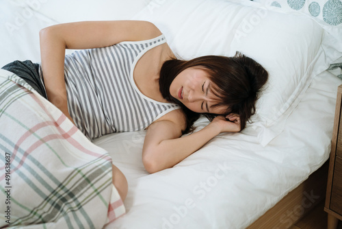 Photos asian lady on her period is lying on bed with painful look since her belly and head hurt