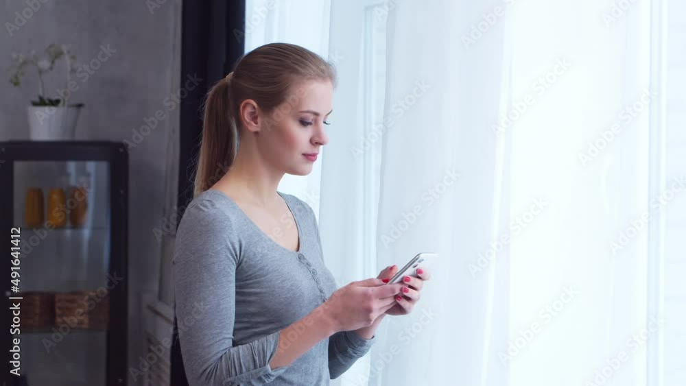 Young teenage girl is standing with a smartphone in front of the window and looking into it. Woman at home in the living room.