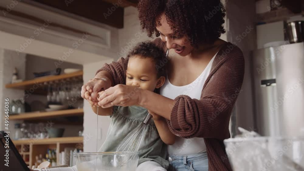 Biracial mother cracking egg in bowl in young daughter's hands in modern-styled kitchen. Baking time with child. Biracial mother cracking egg in bowl in young daughter's hands in modern-styled kitchen. Baking time with child.