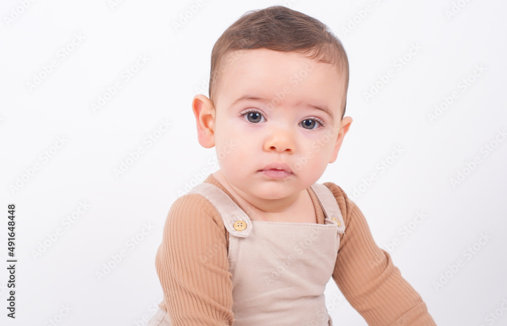Close up portrait of adorable seven month old baby boy wearing beige overalls sitting on white background and looking to the camera front view. Childhood and innocence concept. Close up portrait of adorable seven month old baby boy wearing beige overalls sitting on white background and looking to the camera front view. Childhood and innocence concept.