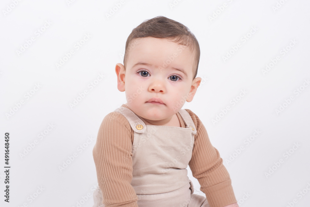 Close up portrait of adorable seven month old baby boy wearing beige overalls sitting on white background and looking to the camera front view. Childhood and innocence concept. 