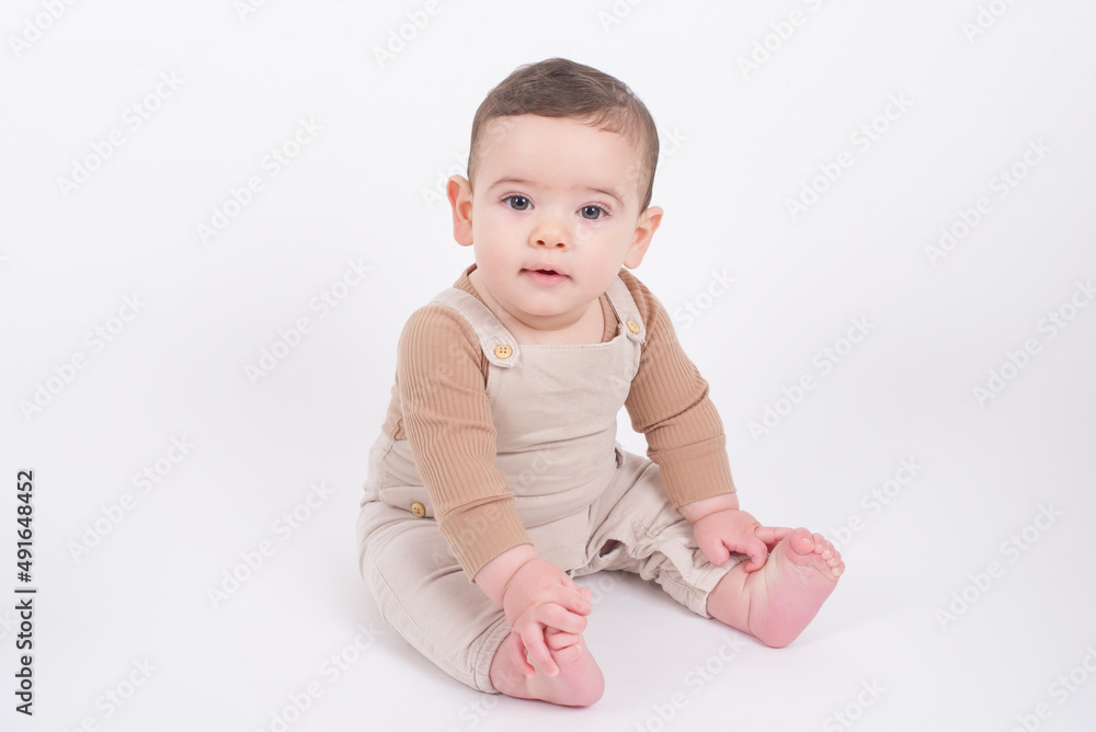 Adorable baby boy wearing beige overalls sitting on white background looking at camera and smiling. Adorable baby boy wearing beige overalls sitting on white background looking at camera and smiling.