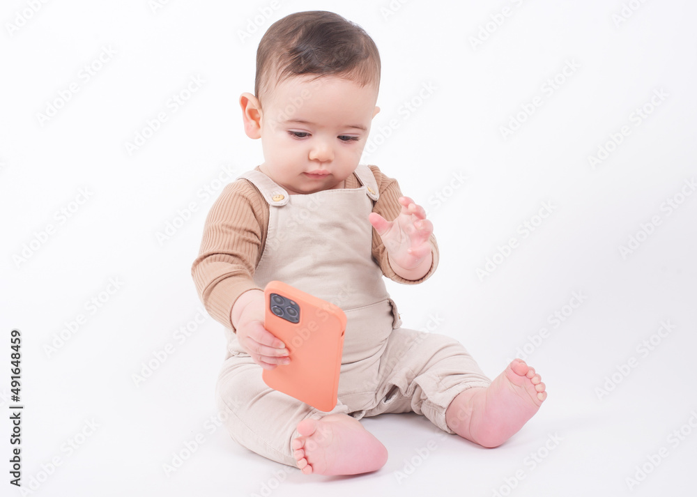 Baby wearing beige overalls sitting on white background holding a telephone and watching cartoons or movies. Baby wearing beige overalls sitting on white background holding a telephone and watching cartoons or movies.