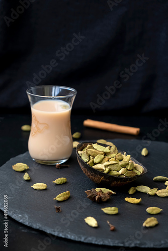 Whole cardamom in coconut shell plate on foreground in focus and traditional oriental karak tea or masala chai on background. Selective focus. Dark background. Exotic cuisine.