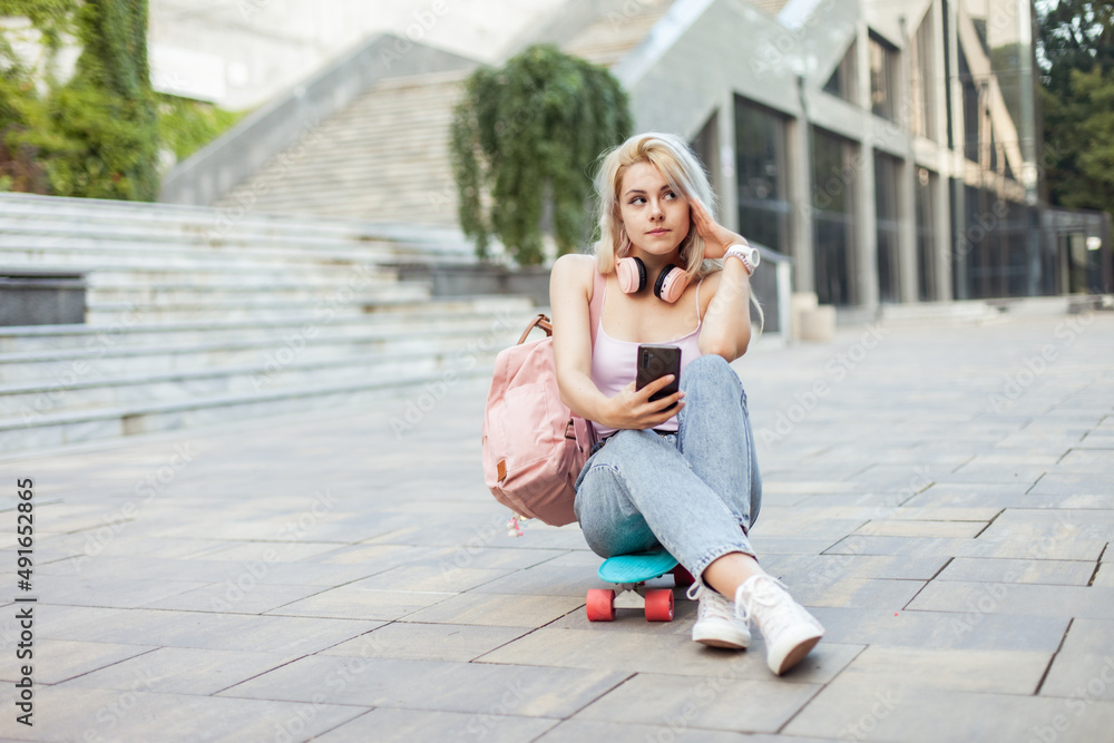 Young smiling cool girl sitting on skateboard and  using phone in the city