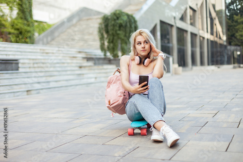 Young smiling cool girl sitting on skateboard and using phone in the city