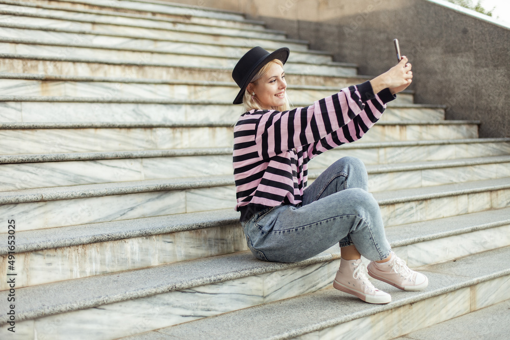 Young charming hipster girl in a hat makes a selfie on the phone while sitting on the stairs outdoors Young charming hipster girl in a hat makes a selfie on the phone while sitting on the stairs outdoors
