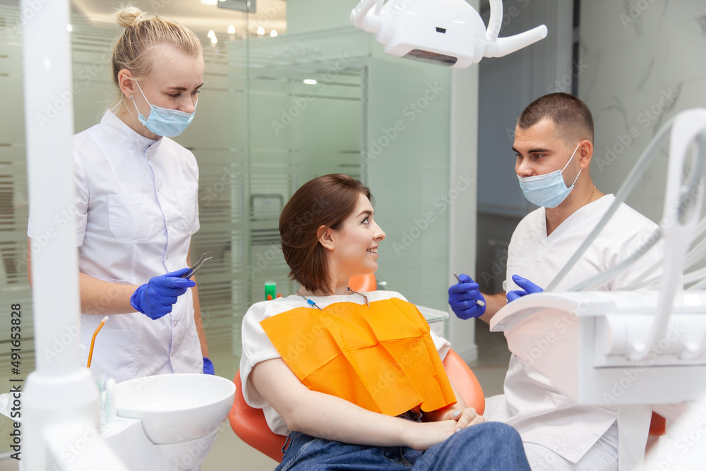 Fototapeta premium Pretty smiling young woman in the dental clinic on examination at dentist. Healthy teeth and beautiful smile concept. Professional doctor with assistant preparing for patient examination