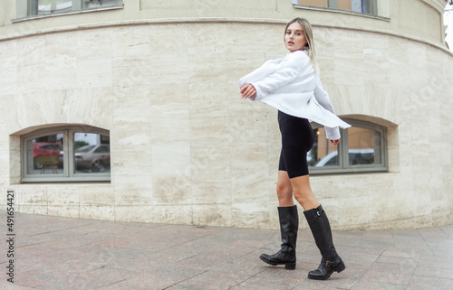 Young fashion blonde woman in white trendy jacket, top and boots posing in city. Street fashion