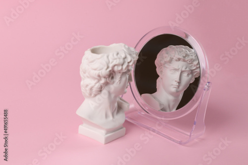 Bust of David admires himself looking in a mirror on a pink background. Narcissism. Minimal still life photo