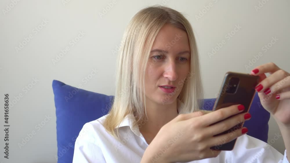 relaxed woman at home communicates via video communication on her smartphone in her bright bedroom or hotel room. She's wearing white shirt and jeans. Online communication. An application or messenger relaxed woman at home communicates via video communication on her smartphone in her bright bedroom or hotel room. She's wearing white shirt and jeans. Online communication. An application or messenger