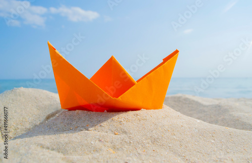 Bright orange paper boat in yellow sand on background of sea waves and blue sky on sunny summer day. Colored ship close-up. Concept adventures travelling vacation holiday rest tourism voyager dream
