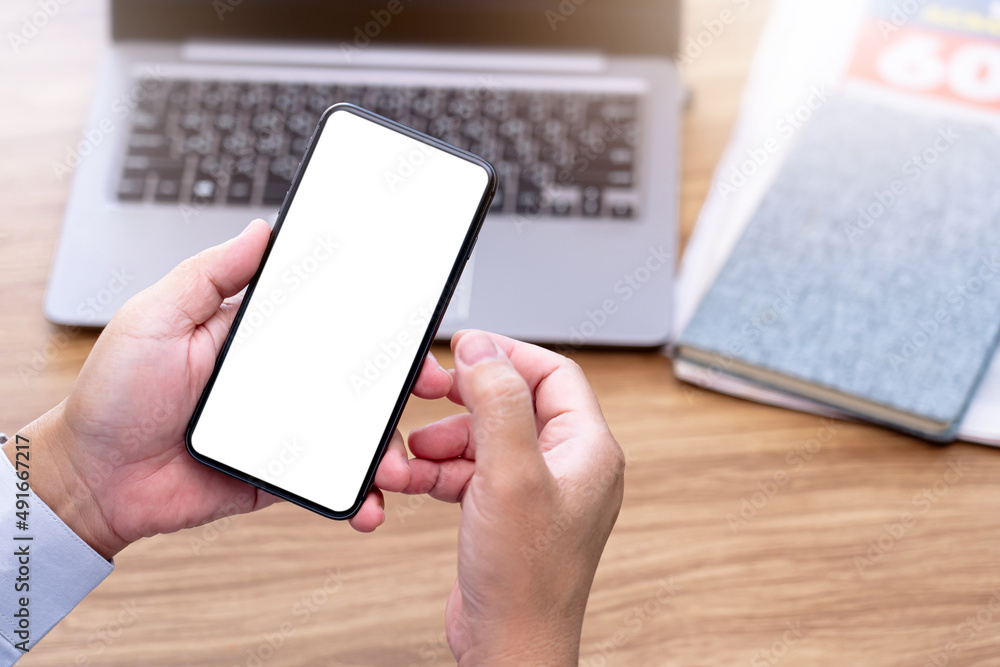 Smartphone in hand a man and a blank white screen, laptop newspaper on wood table, business concept Smartphone in hand a man and a blank white screen, laptop newspaper on wood table, business concept
