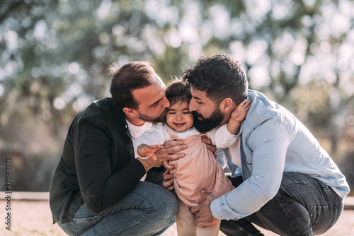 Gay couple kissing their daughter who is smiling amusedly in an urban park.