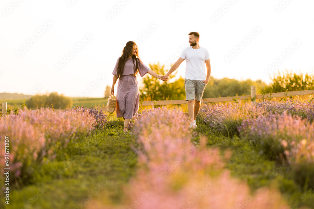 Stylish young adult couple walking at lavender field, having a romantic date. Handsome man hold a hand of beautiful woman, enjoy moments together, meeting the sunset, spend weekends outdoors concept