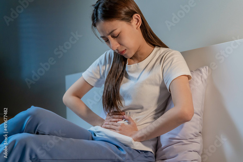 Flatulence ulcer, asian young woman in belly, stomach ache from food poisoning, abdominal pain and digestive problem, gastritis or diarrhoea sitting in bed room in the night. Abdomen inflammation. photo