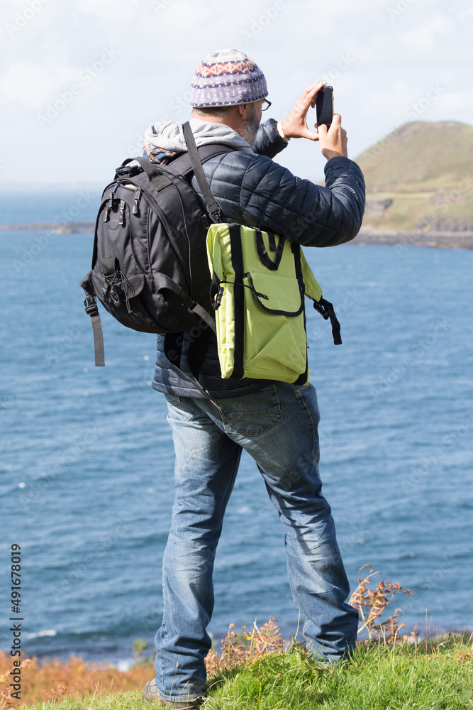 A man stands taking a photo with a mobile phone.he has rucksack on his back and a green folding chair mat