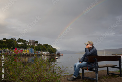 A man sits on a bench making a phone call on a mobile phone.He wears a baseball hat.In background is sea and coloured buildings of Tobermory in Scotland photo