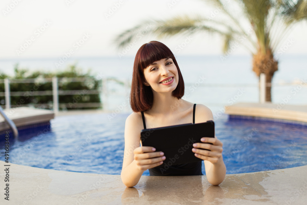 Happy smiling girl using digital tablet computer while bathing in a swimming pool with blue water in summer vacations at luxury sea resort