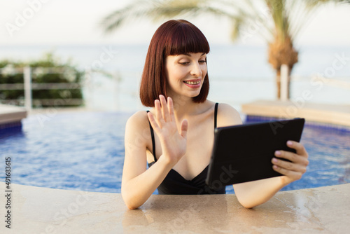 Happy smiling young woman holding digital tablet at outdoors swimming pool, having a video call, waving to her friend. Vacation, gadgets and technology concept