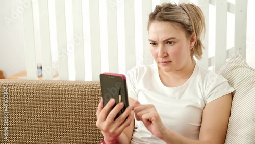 Caucasian woman is sitting at home on couch, looking at smartphone screen and frowning. Reads bad news, gets upset. Concept of mental burnout and doom scrolling.