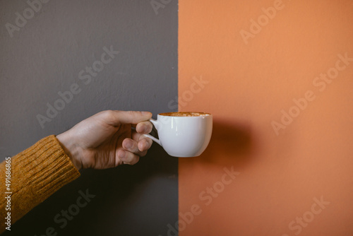A man's hand holding a white coffee mug against the background of a wall painted in two colors - orange and gray.