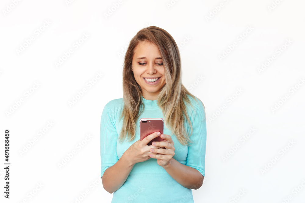 A young caucasian happy pretty blonde woman smiles and holds a mobile phone looking at the screen isolated on a white background. The girl in a turquoise blouse uses her smartphone A young caucasian happy pretty blonde woman smiles and holds a mobile phone looking at the screen isolated on a white background. The girl in a turquoise blouse uses her smartphone
