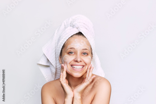 A young caucasian happy smiling woman with a white towel on her head after a shower applies a moisturizing cream isolated on a white background. Skin care, cosmetology. Morning routine. Close up