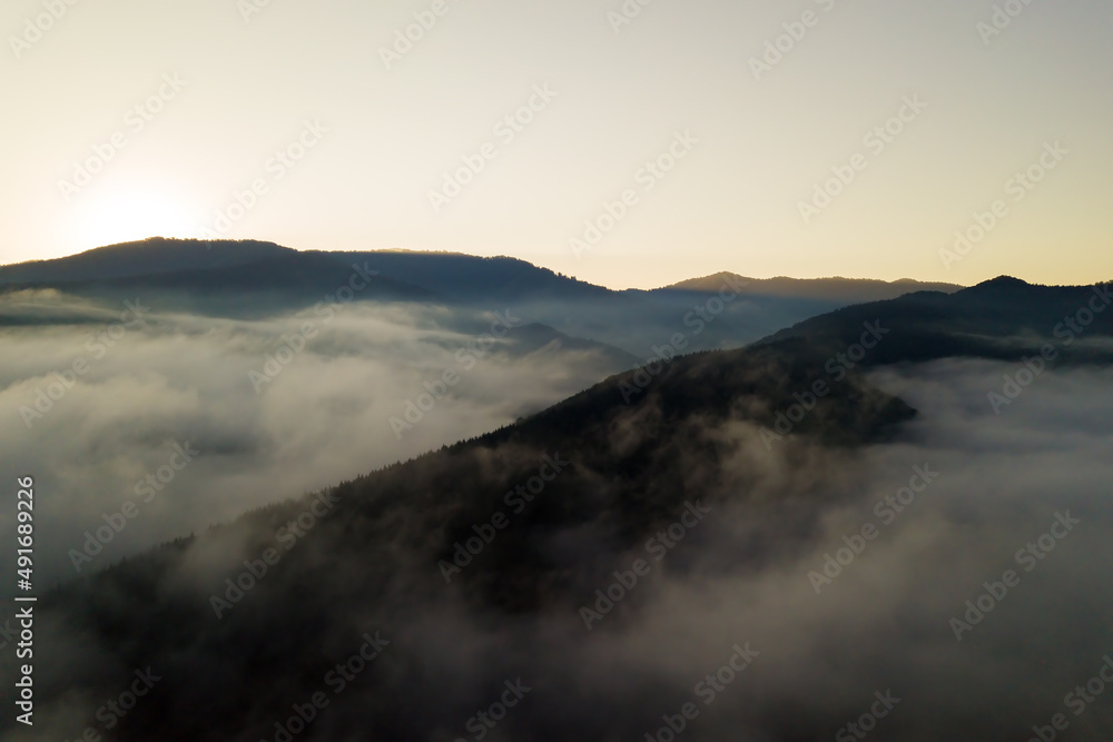 Aerial view of vibrant sunrise over white dense fog with distant dark silhouettes of mountain hills on horizon Aerial view of vibrant sunrise over white dense fog with distant dark silhouettes of mountain hills on horizon
