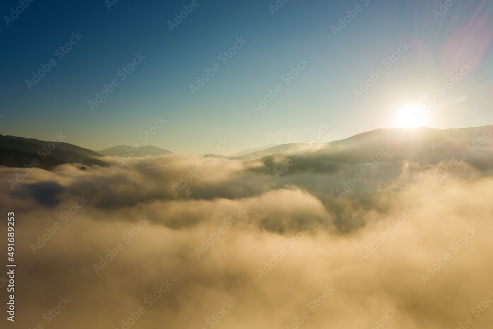 Aerial view of vibrant sunrise over white dense fog with distant dark silhouettes of mountain hills on horizon Aerial view of vibrant sunrise over white dense fog with distant dark silhouettes of mountain hills on horizon