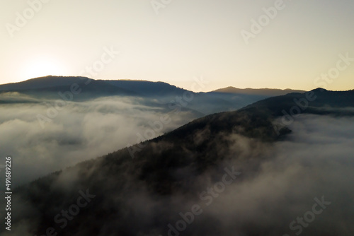 Aerial view of vibrant sunrise over white dense fog with distant dark silhouettes of mountain hills on horizon