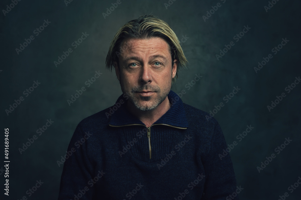 Rugged blond man with a stubble in a blue woolen sweater with zipper collar in front of a dark grey wall. Rugged blond man with a stubble in a blue woolen sweater with zipper collar in front of a dark grey wall.