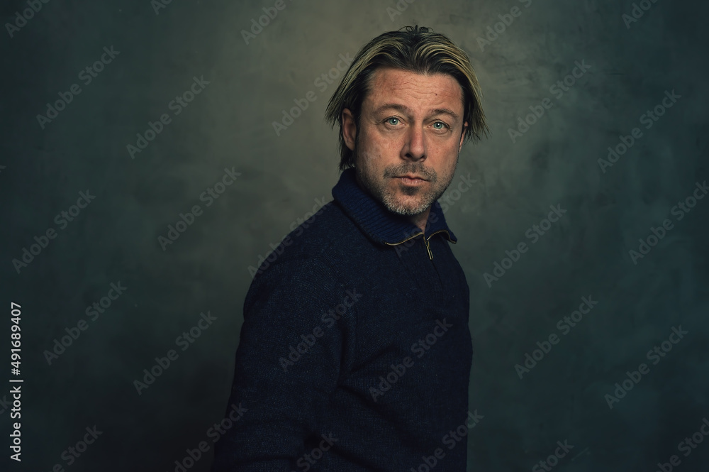 Rugged blond man with a stubble in a blue woolen sweater with zipper collar in front of a dark grey wall. Rugged blond man with a stubble in a blue woolen sweater with zipper collar in front of a dark grey wall.