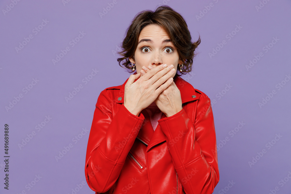 Young shocked surprised amazed scared astonished woman 20s wearing red leather jacket cover mouth with hand isolated on plain pastel light purple background studio portrait. People lifestyle concept. Young shocked surprised amazed scared astonished woman 20s wearing red leather jacket cover mouth with hand isolated on plain pastel light purple background studio portrait. People lifestyle concept.