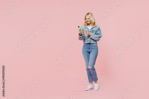 Full size elderly smiling fun happy woman 50s wearing denim jacket hold in hand use mobile cell phone typing message isolated on plain pastel light pink background. People lifestyle fashion concept.