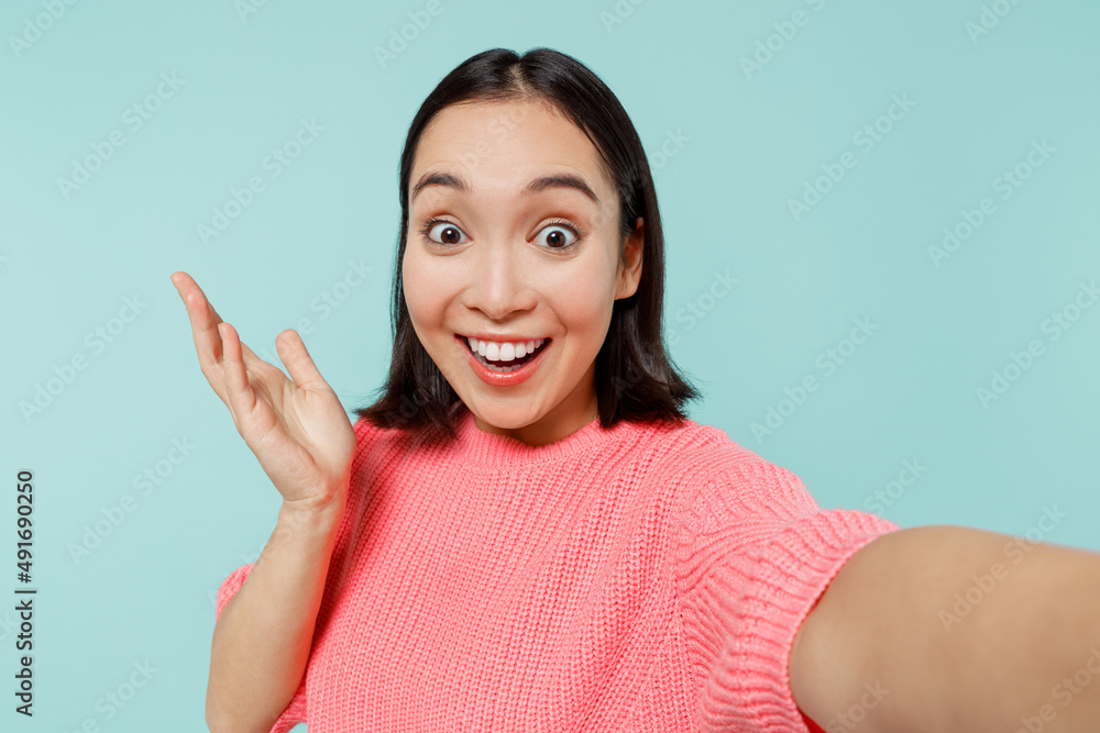 Close up young happy fun woman of Asian ethnicity 20s in pink sweater do selfie shot pov on mobile phone spread hands isolated on pastel plain light blue background studio. People lifestyle concept. Close up young happy fun woman of Asian ethnicity 20s in pink sweater do selfie shot pov on mobile phone spread hands isolated on pastel plain light blue background studio. People lifestyle concept.