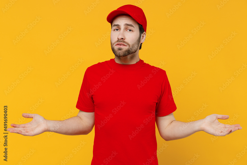 Professional confused delivery guy employee man in red cap T-shirt uniform workwear work as dealer courier spread hands shrugging shoulders isolated on plain yellow background studio. Service concept.