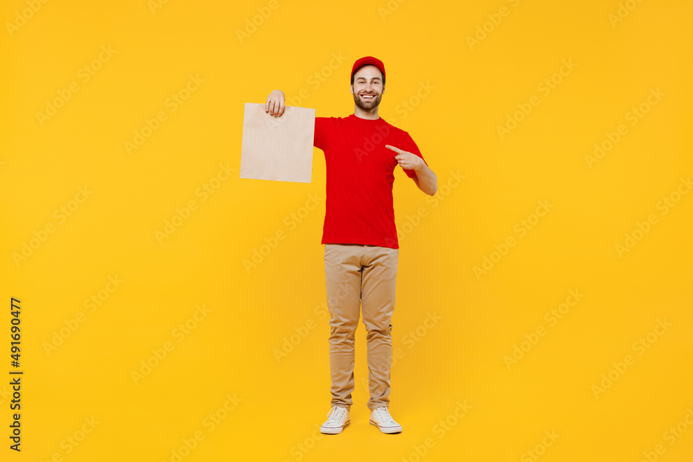 Full body delivery guy employee man in red cap T-shirt uniform workwear work as dealer courier point on brown clear blank craft paper takeaway bag mock up isolated on plain yellow background studio Full body delivery guy employee man in red cap T-shirt uniform workwear work as dealer courier point on brown clear blank craft paper takeaway bag mock up isolated on plain yellow background studio