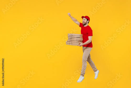Side view full body delivery guy employee man in red cap T-shirt uniform workwear work as dealer courier hold pizza in cardboard flatbox jump high isolated on plain yellow background Service concept