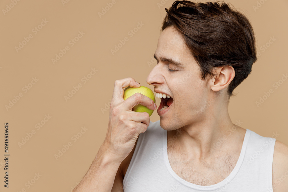 Close up fun attractive caucasian young man 20s perfect skin in undershirt hold bite green apple isolated on pastel pastel beige background studio. Skin care healthcare cosmetic procedures concept Close up fun attractive caucasian young man 20s perfect skin in undershirt hold bite green apple isolated on pastel pastel beige background studio. Skin care healthcare cosmetic procedures concept