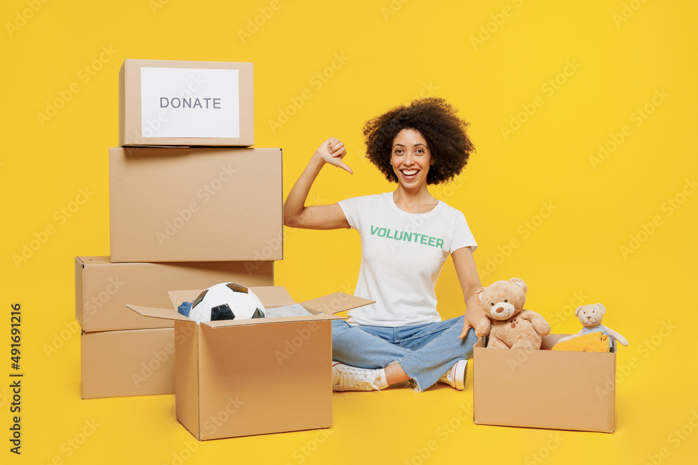 Full size young woman of African American ethnicity in white volunteer t-shirt hold boxes with presents point fingers on herself isolated on plain yellow background. Voluntary free work help concept. Full size young woman of African American ethnicity in white volunteer t-shirt hold boxes with presents point fingers on herself isolated on plain yellow background. Voluntary free work help concept.