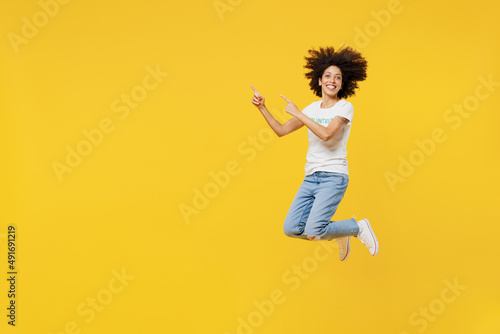 Full body young woman of African American ethnicity wears white volunteer t-shirt jump high point finger aside isolated on plain yellow background. Voluntary free work assistance help grace concept. photo