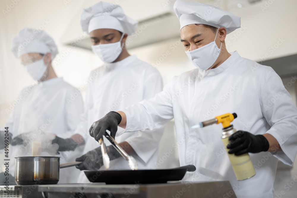 Multiracial team of chef cooks in white uniform cooking together in the kitchen. Asian chef frying meat with burner, latin and european guy cooking on background. Cooks wearing face masks and