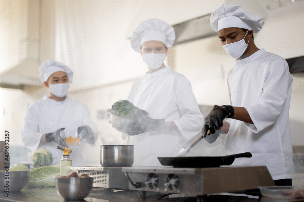 Multiracial team of cooks in uniform and face masks cooking meals for a restaurant in the kitchen. Concept of teamwork at restaurant during pandemic. Latin, Asian and European guys cooking together